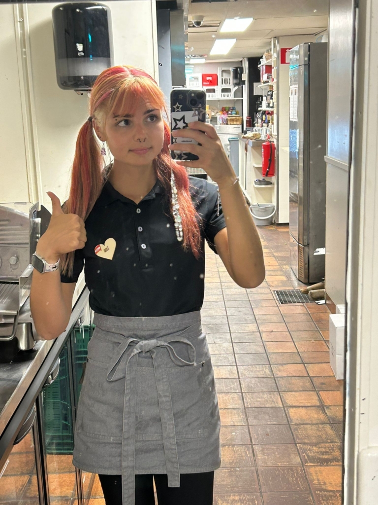 Eine junge Frau macht in Arbeitskleidung ein Spiegelselfie in einem Hotelbetrieb.