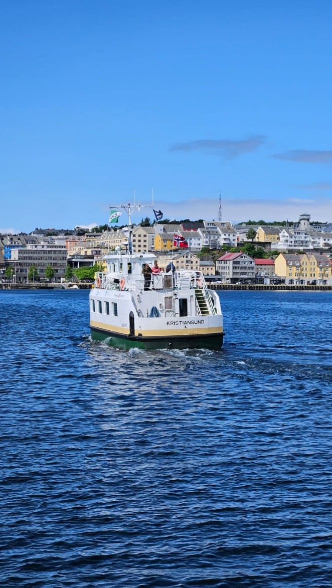 Eine kleine Fähre fährt vor den bunten Häusern von Kristiansund über das Wasser.