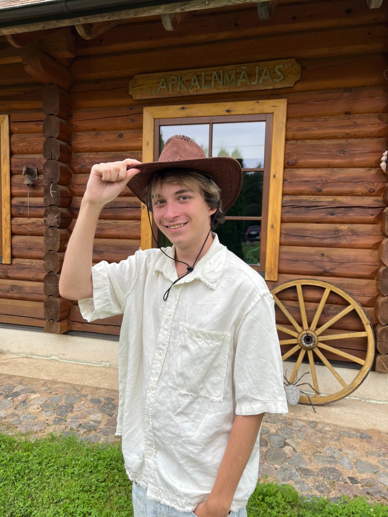 Ein junger Mann trägt einen braunen Hut und steht vor einem Holzhaus mit einem Wagenrad.